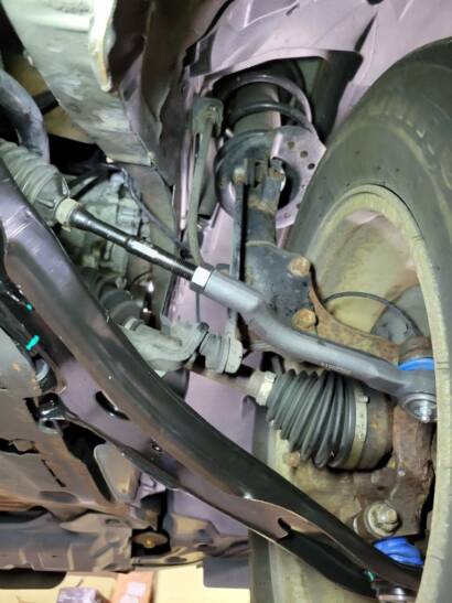 Mechanic's ratchet on the mounting bolt of a new lower control arm, seen from under the car with the coil spring and CV axle in view